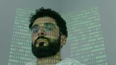 A man with a beard and eyeglasses looking at the camera, illuminated by binary code projection.