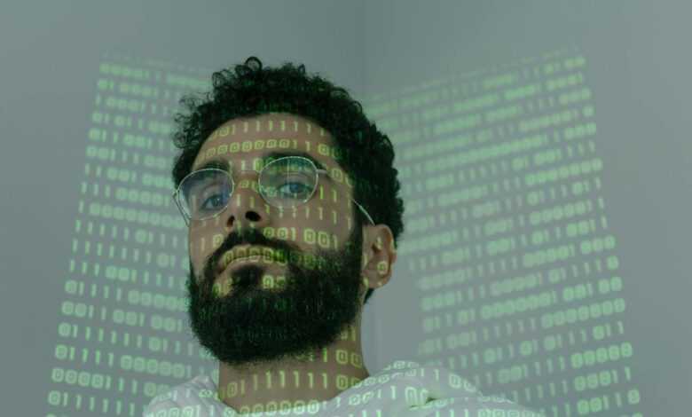 A man with a beard and eyeglasses looking at the camera, illuminated by binary code projection.